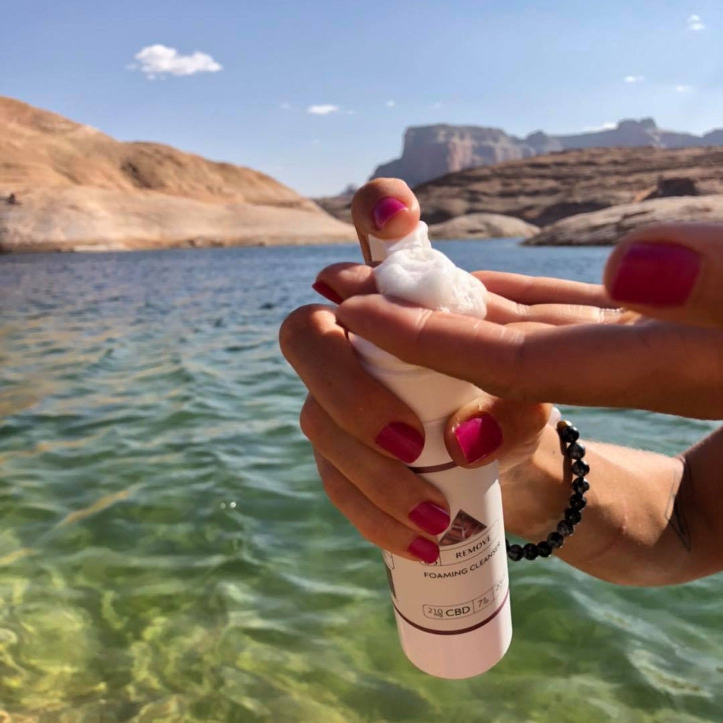 Person holding Color Up REMOVE Foaming Cleanser in 5 oz retail size with scenic background of water and mountains.