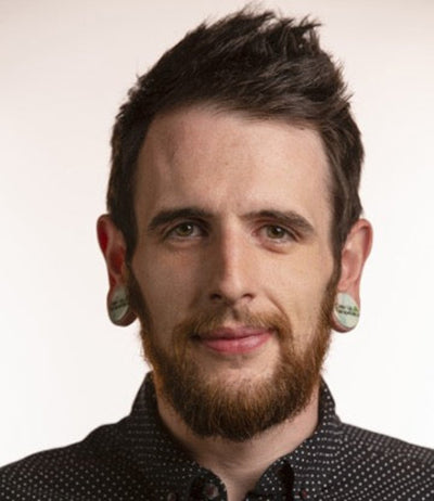 Man with a beard and earrings wearing a patterned shirt against a plain background