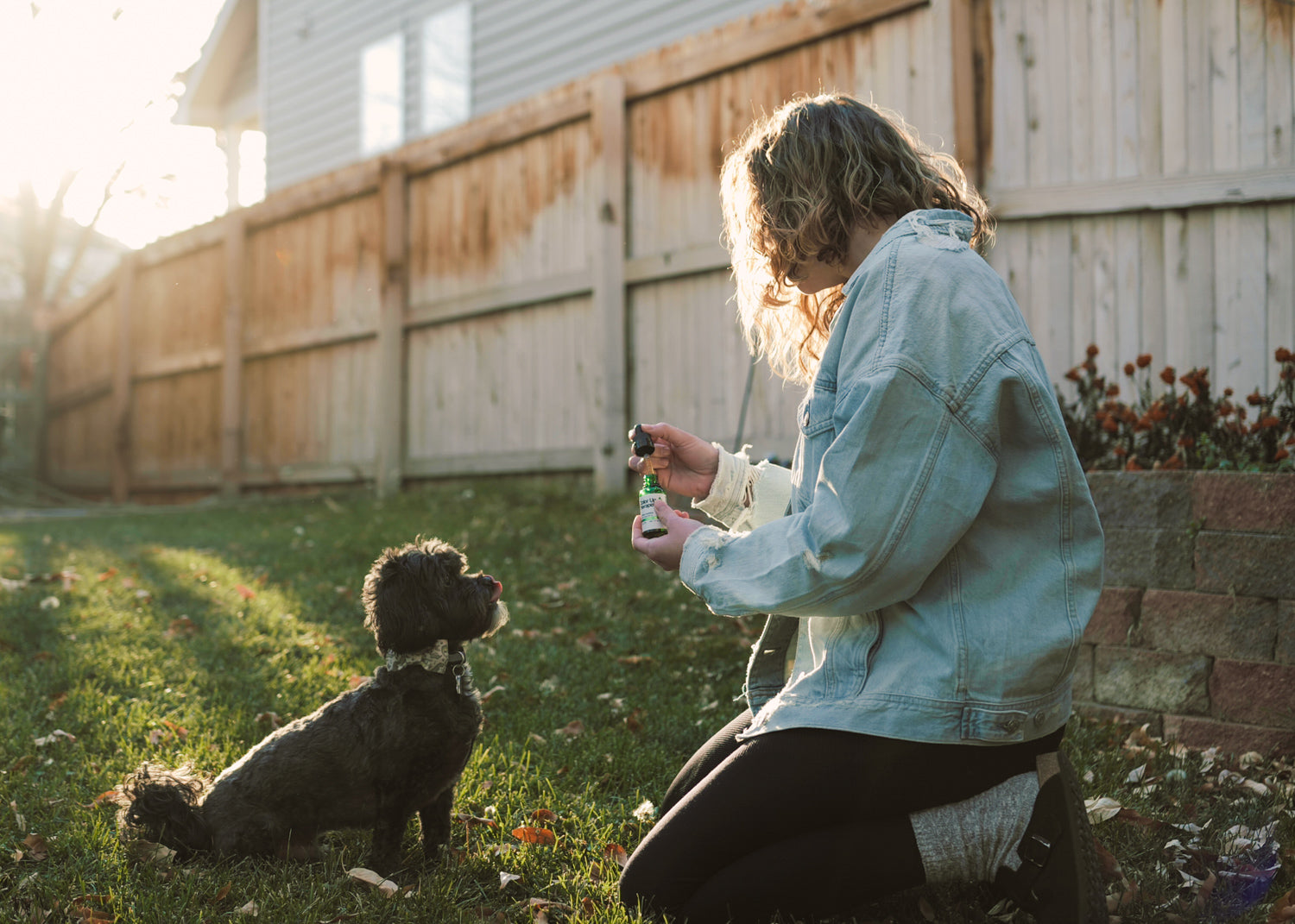 Person kneeling in a backyard with a small black dog, holding a bottle.