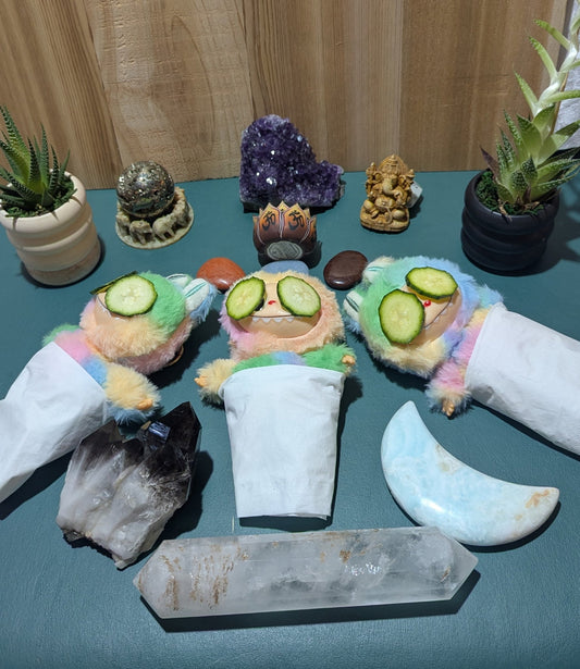 Three Labubu dolls laying down in a semi-circle in a scene setup like a massage and skincare room with cucumbers on their eyes, and crystals in the background