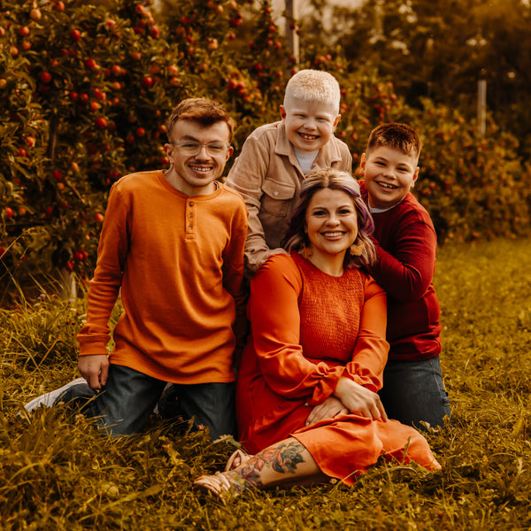 A woman, Amanda, educator for Color Up CBD Skincare, with her three boys around her sitting on the grass with fruit trees in the background.