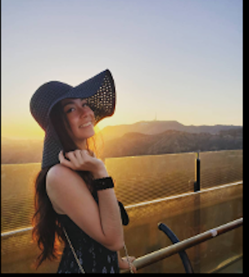 Amber, co-owner of Color Up with a black hat on, in front of mountain range with sun shining in the background.