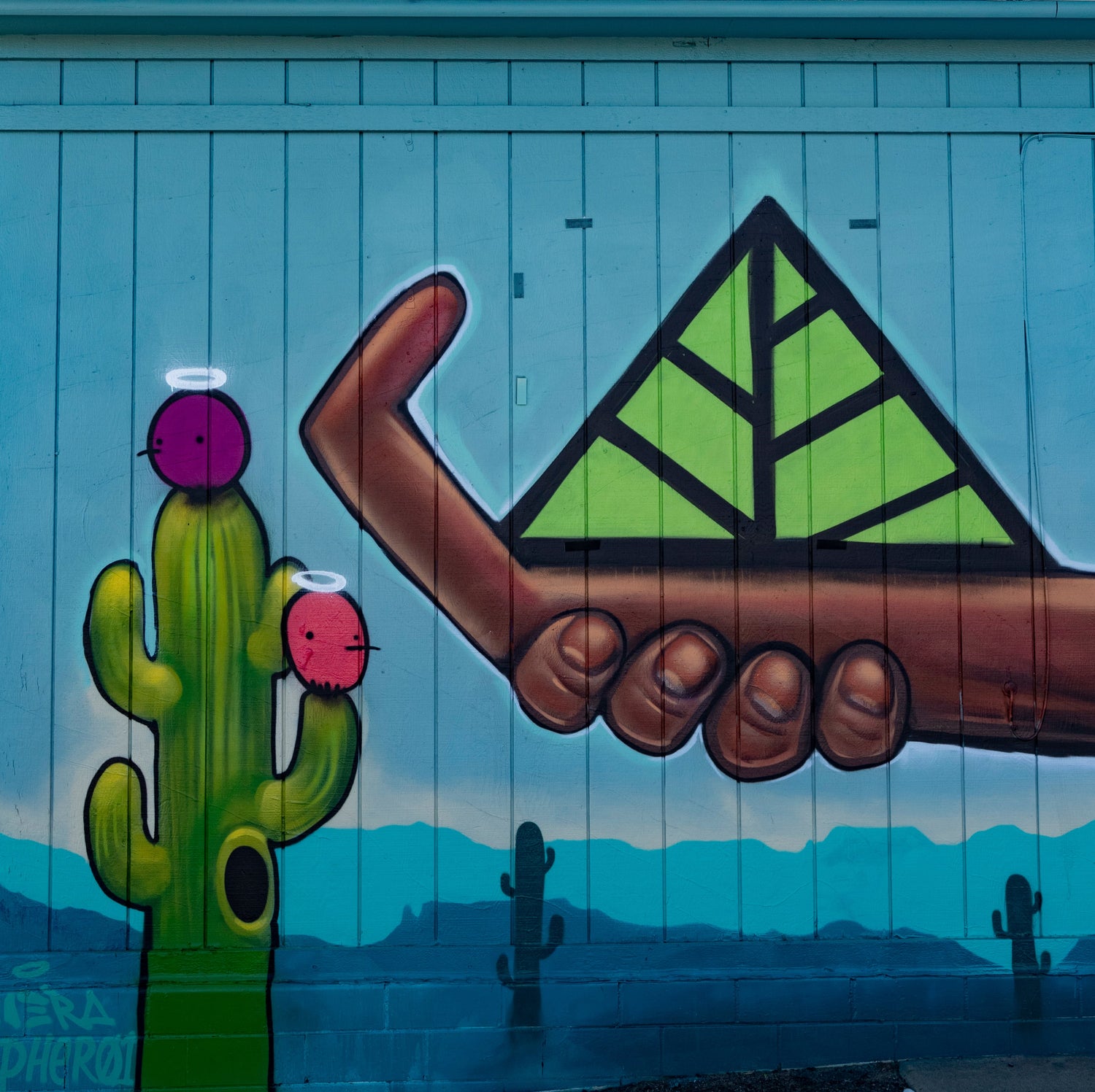 Mural of a hand holding Color Up's green leaf logo above a cactus with a blue background on Color Up Wellness Center building in 2020.
