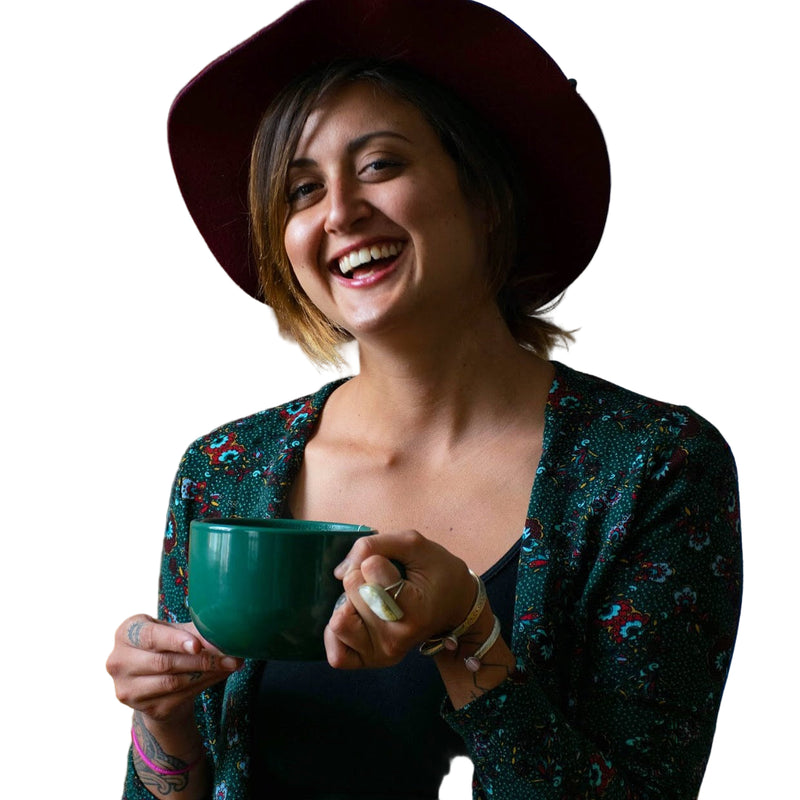 Woman wearing a wide-brimmed hat and floral cardigan, holding a green mug in a cozy indoor setting.