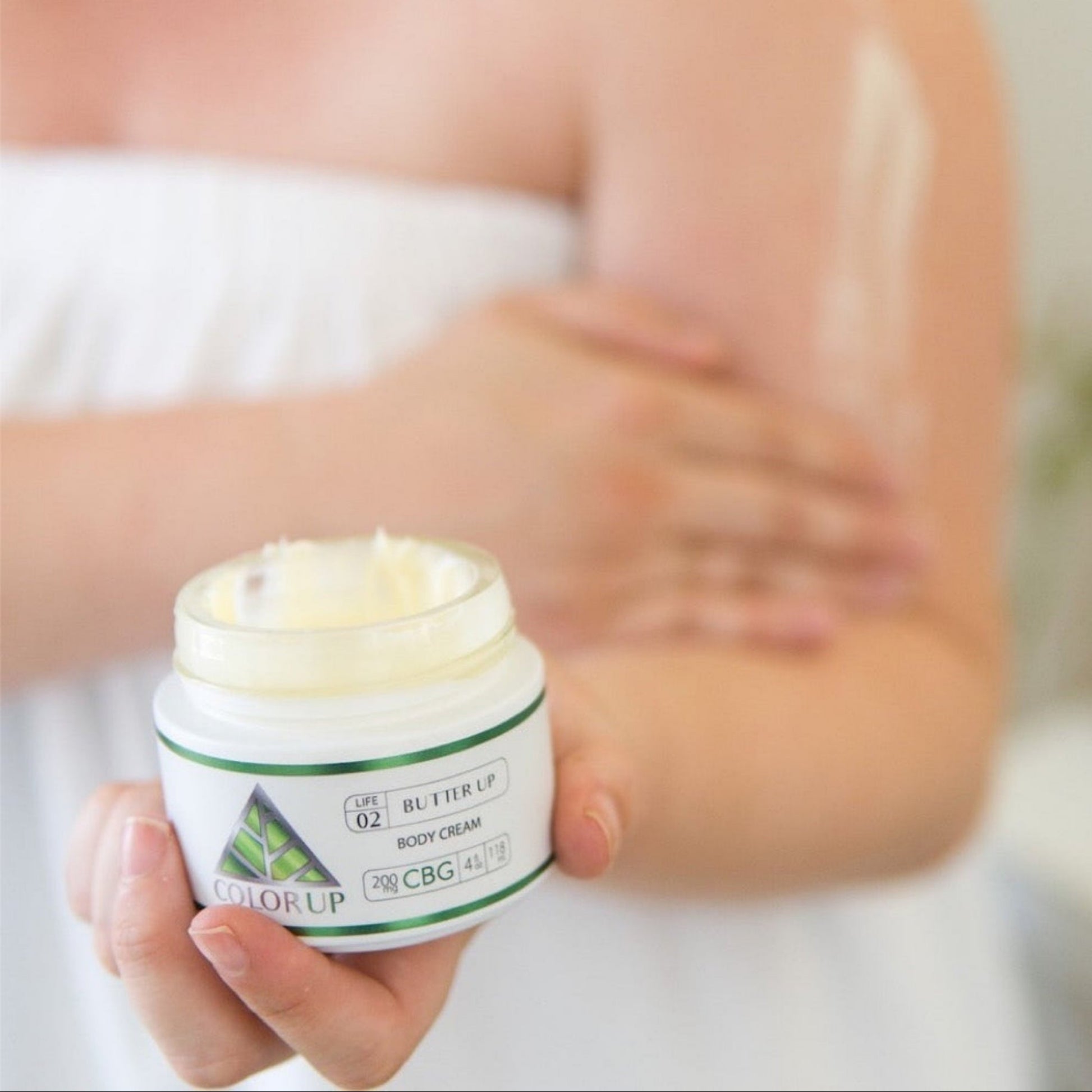 Hand holding jar with top off and cream being applied to body – Color Up label with green triangle on jar. 