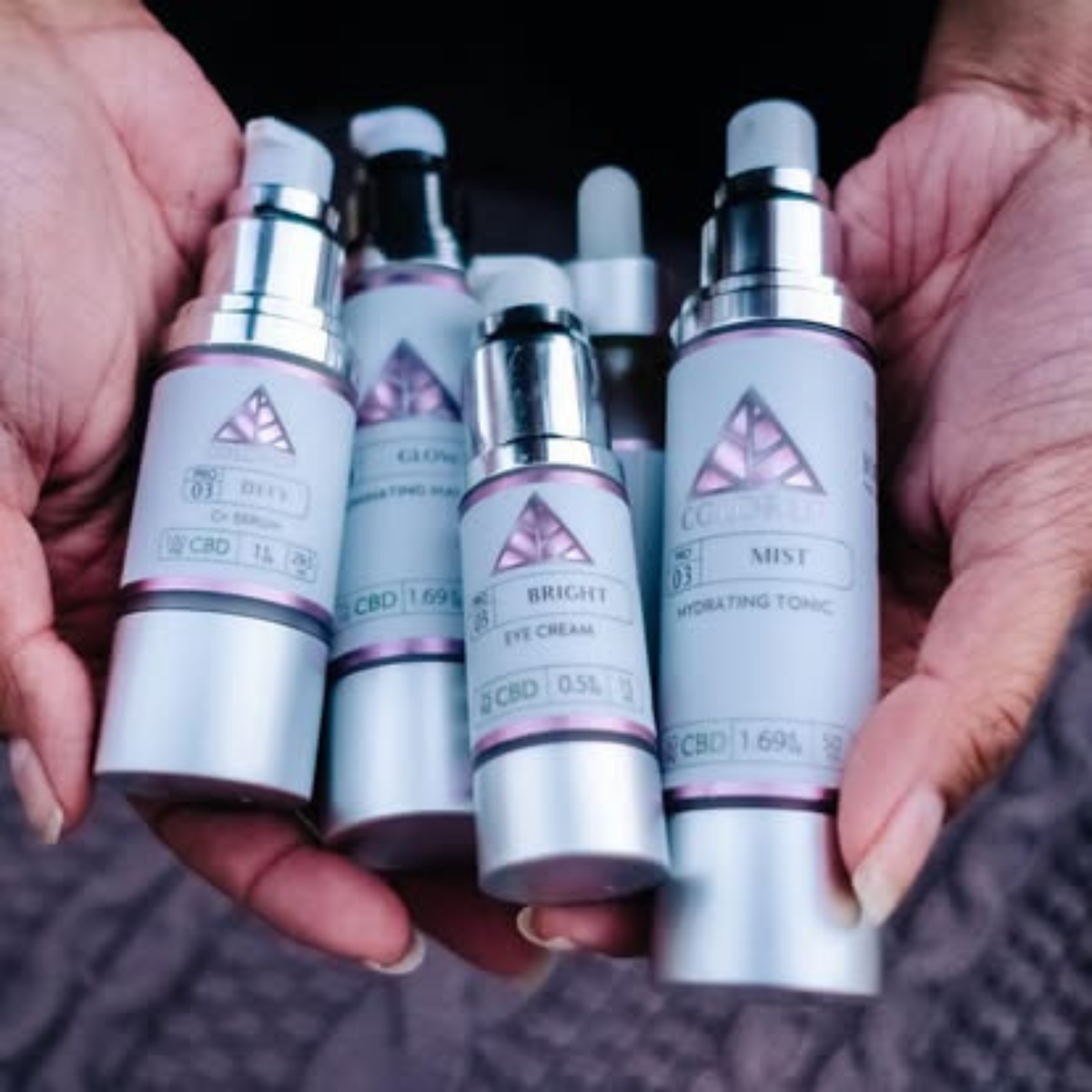Four Color Up PRO cosmetic bottles with chrome and white labels are held in two hands against a dark textured background. The labels feature a pink geometric logo and text including product names. 