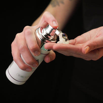 Close-up of hands holding a white 'Color Up Remove Foaming Cleanser' bottle against a black background