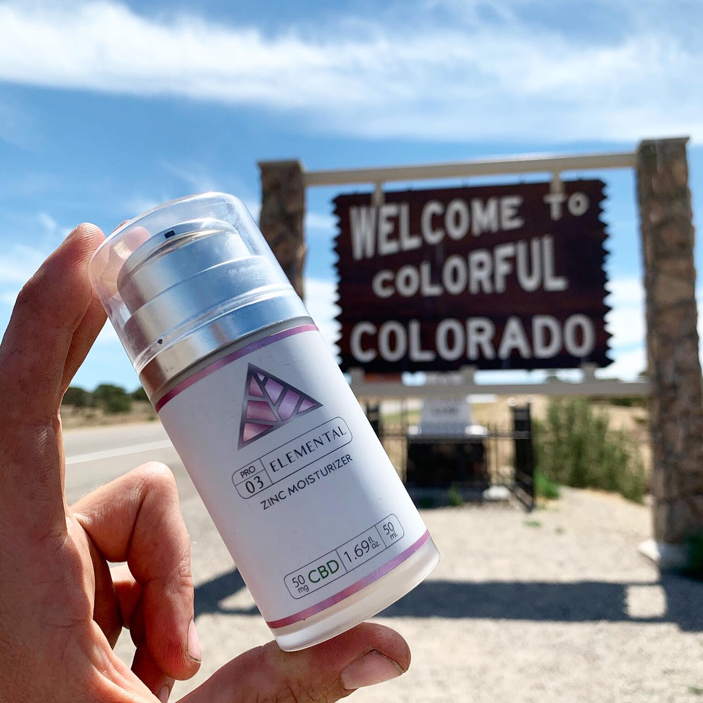 Hand holding a silver cosmetic bottle with 'color up elemental zinc moisturizer' and pink logo on white label in front of 'Welcome to Colorful Colorado' sign.